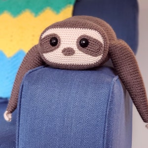 Pode incluir: Um brinquedo de preguiça de crochê com pelo marrom e rosto bege. A preguiça está sentada em um sofá azul com um padrão de espinha de peixe.