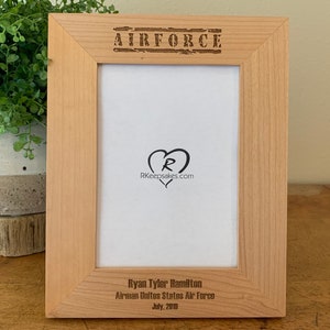 May include: A wooden picture frame with the words "AIRFORCE" engraved on the top. The frame has a white mat and is ready to hold a photo. The bottom of the frame has the text "Ryan Tyler Hamilton Airman United States Air Force July, 2018" engraved on it.