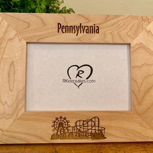 May include: A wooden picture frame with an engraved design of a Ferris wheel and roller coaster. The frame is light brown and has the text "Pennsylvania" and "RKeepsakes.com" engraved on it.