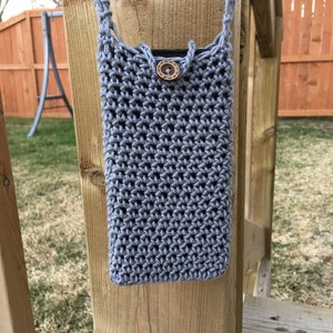 May include: A light grey, crocheted phone case with a strap. The case is rectangular with a button closure. The button is a light brown, wooden circle. The case is hanging on a wooden post.