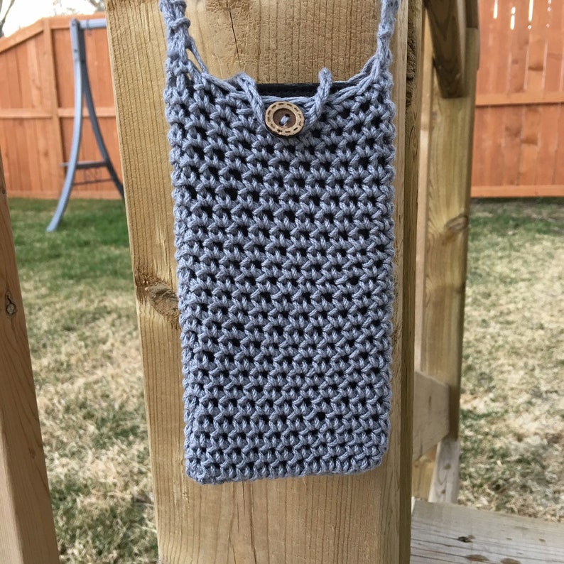 May include: A light grey, crocheted phone case with a strap. The case is rectangular with a button closure. The button is a light brown, wooden circle. The case is hanging on a wooden post.