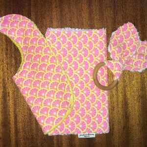 May include: A pink and yellow baby burp cloth, a pink and yellow baby teether, and a wooden teether ring. The fabric has a pattern of pink and yellow citrus slices.