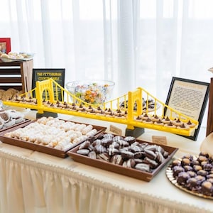 May include: A dessert table featuring a yellow bridge-shaped display filled with cookies. Various trays hold an assortment of cookies, including chocolate-covered, iced, and sandwich cookies. A sign reads "The Pittsburgh Cookie Table".