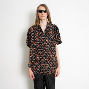 Może przedstawiać: Czarna koszula z krótkim rękawem zapinana na guziki z brązowym i białym geometrycznym wzorem. Koszula ma kołnierzyk i luźny krój.