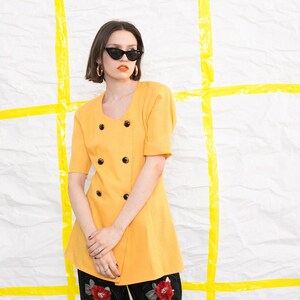 May include: A woman wearing a yellow double-breasted jacket with black buttons and black cat-eye sunglasses. She is standing in front of a white background with yellow tape.