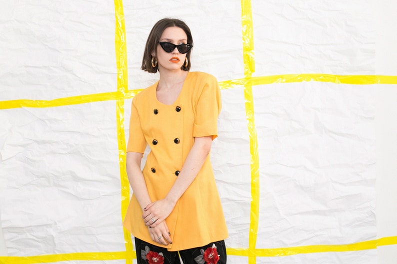 May include: A woman wearing a yellow double-breasted jacket with black buttons and black cat-eye sunglasses. She is standing in front of a white background with yellow tape.