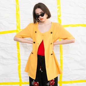 May include: A yellow short-sleeved jacket with a double-breasted closure and a red top underneath. The jacket has a slightly flared hem and is worn by a person with dark hair and sunglasses.