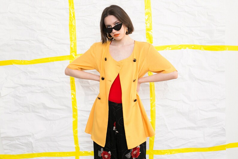 May include: A yellow short-sleeved jacket with a double-breasted closure and a red top underneath. The jacket has a slightly flared hem and is worn by a person with dark hair and sunglasses.
