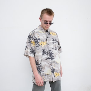 Könnte beinhalten: Ein kurzärmeliges Hemd mit Knöpfen und einem tropischen Druck mit Palmen, Blumen und einer Strandszene. Das Hemd ist beige mit schwarzen und gelben Akzenten.