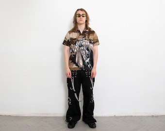 May include: A person wearing a brown and tan short-sleeved shirt with a graphic print of a person with a sword. The person is wearing black pants with silver chains and black shoes.