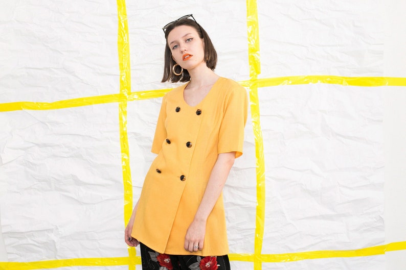 May include: A yellow double-breasted tunic with short sleeves and black buttons. The tunic is worn by a person with short brown hair and black sunglasses.