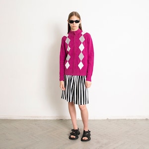 May include: A woman wearing a pink cardigan with a white diamond pattern, a black and white striped skirt, and black sandals.