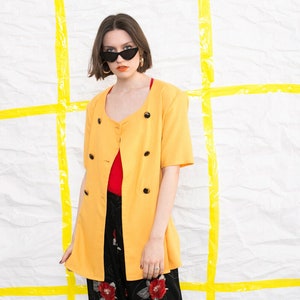 May include: A yellow blazer with a double-breasted closure and short sleeves. The blazer has black buttons and is worn over a red top and black trousers with a floral print.