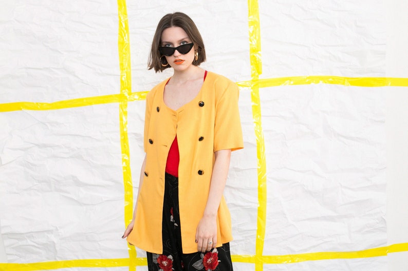 May include: A yellow blazer with a double-breasted closure and short sleeves. The blazer has black buttons and is worn over a red top and black trousers with a floral print.