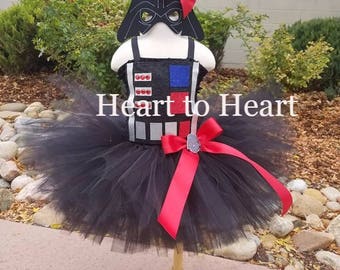Può includere: Gonna tutu in tulle nero con un nastro rosso e un top da costume Darth Vader nero e bianco. Il top del costume ha accenti rossi e blu e una maschera nera. Il testo "Heart to Heart" è visibile nell'immagine.