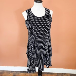May include: A sleeveless black dress with white polka dots, displayed on a mannequin. The dress features a scoop neckline and a tiered, ruffled skirt. The mannequin is on a black stand, set against a peach-colored wall.
