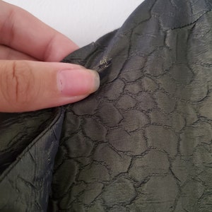 May include: Close-up of a dark green fabric with a textured pattern of small circles. The fabric is slightly wrinkled and has a small hole in the upper right corner.
