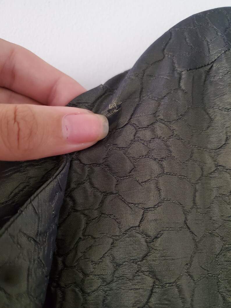 May include: Close-up of a dark green fabric with a textured pattern of small circles. The fabric is slightly wrinkled and has a small hole in the upper right corner.