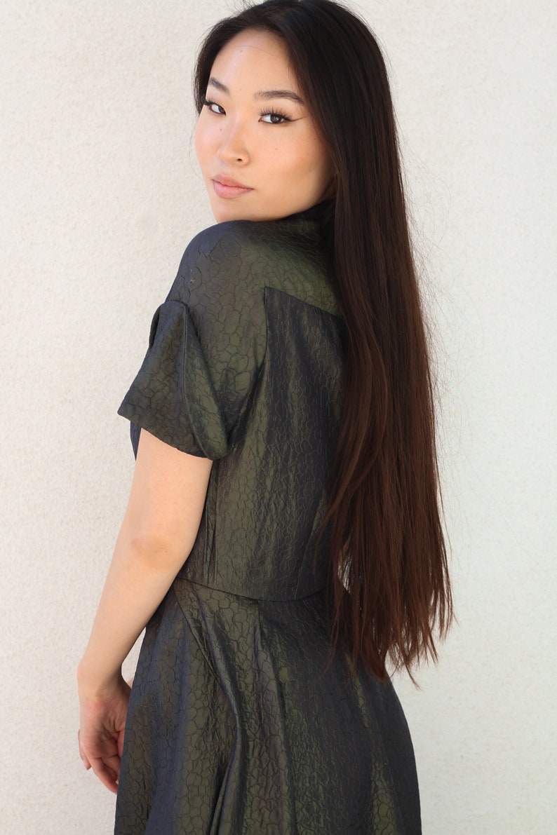 May include: A woman with long dark hair is wearing a dark green, textured, short-sleeved dress. She is looking over her shoulder.