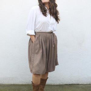 May include: A woman wearing a white button-down shirt and a brown pleated skirt with a button closure. She is wearing brown leather boots.