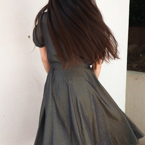 May include: A woman wearing a dark green, textured, knee-length dress with a fitted bodice and a full skirt. Her long, dark hair is flowing behind her.
