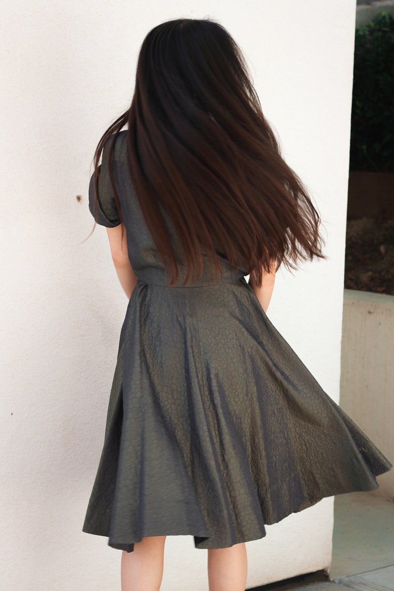 May include: A woman wearing a dark green, textured, knee-length dress with a fitted bodice and a full skirt. Her long, dark hair is flowing behind her.