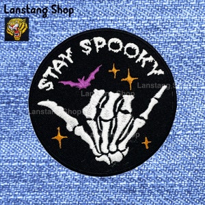 May include: Black and white embroidered patch with a skeleton hand giving the shaka sign. The patch has the text "STAY SPOOKY" in white lettering around the edge. There are three small yellow stars and a purple bat in the center of the patch.