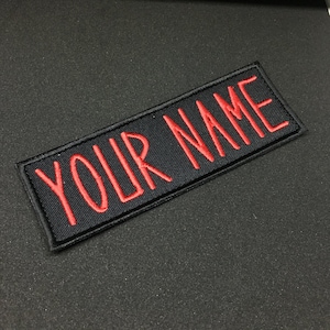 May include: Black fabric patch with red embroidered text that reads "YOUR NAME".