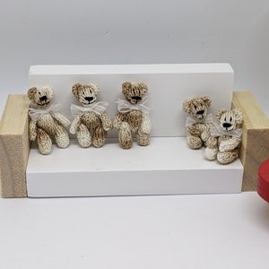 May include: Five small knitted teddy bears with white bows sitting on a white shelf. The bears are brown and white and are arranged in a row.