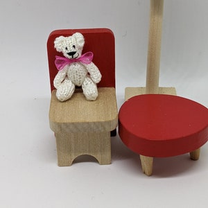 May include: A miniature scene with a white crocheted teddy bear wearing a pink bow, sitting on a small wooden chair with a red back. A red table and a green lamp are also present, all on a white surface.