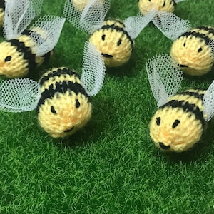 May include: Handmade crocheted bees with yellow and black striped bodies and white tulle wings. The bees are arranged on a green, grassy surface. Each bee has a smiling face, creating a whimsical and playful scene.