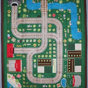Puede incluir: Una alfombra de juego de fieltro verde con un diseño de vía de tren. La vía es un bucle con un túnel, una estación y una señal de stop. También hay pequeños jardines y un estanque en la alfombra.