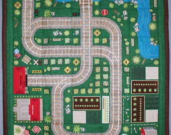 May include: A green felt play mat with a train track design. The track is a loop with a tunnel, a station, and a stop sign. There are also small gardens and a pond on the mat.