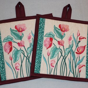 Puede incluir: Dos agarraderas de olla acolchadas con un diseño floral. Las agarraderas son de color burdeos con un patrón floral verde y blanco. Las flores son rosas y blancas.