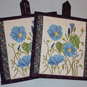 May include: Two purple and white quilted pot holders with a floral design featuring blue, green, and yellow embroidered morning glory flowers.
