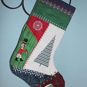 May include: A green, red, white, and blue patchwork Christmas stocking with an embroidered nutcracker, Christmas tree, and drum.