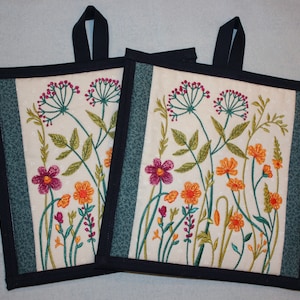 May include: Two cream-coloured pot holders with a floral embroidery design. The pot holders have a dark blue border and a dark blue hanging loop.