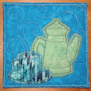 May include: A blue quilted coaster with a green teapot and a blue and white teacup with a saucer. The teapot and teacup are embroidered with a swirling design.