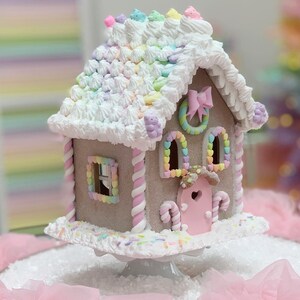 K&ouml;nnte beinhalten: Ein skurriles Lebkuchenhaus mit einem pastellfarbenen Regenbogen-Dach, wei&szlig;em Zuckerguss und rosa-wei&szlig;en Zuckerstangen-Akzenten. Das Haus hat herzf&ouml;rmige Fenster und eine rosa T&uuml;r mit einem Herz-Ausschnitt. Es steht auf einem wei&szlig;en Sockel.