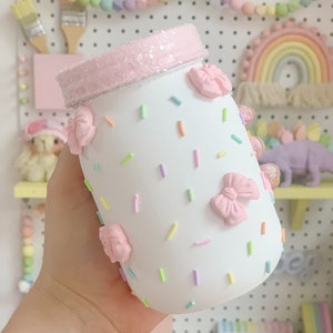 May include: A white glass jar decorated with pastel-colored sprinkles and three-dimensional pink bows. The rim of the jar is coated in a textured, light pink glitter finish. The jar features a smooth, matte white surface with scattered sprinkles.