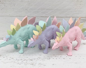 May include: Three pastel-colored dinosaur figurines with spiky backs. The dinosaurs are green, purple, and pink.