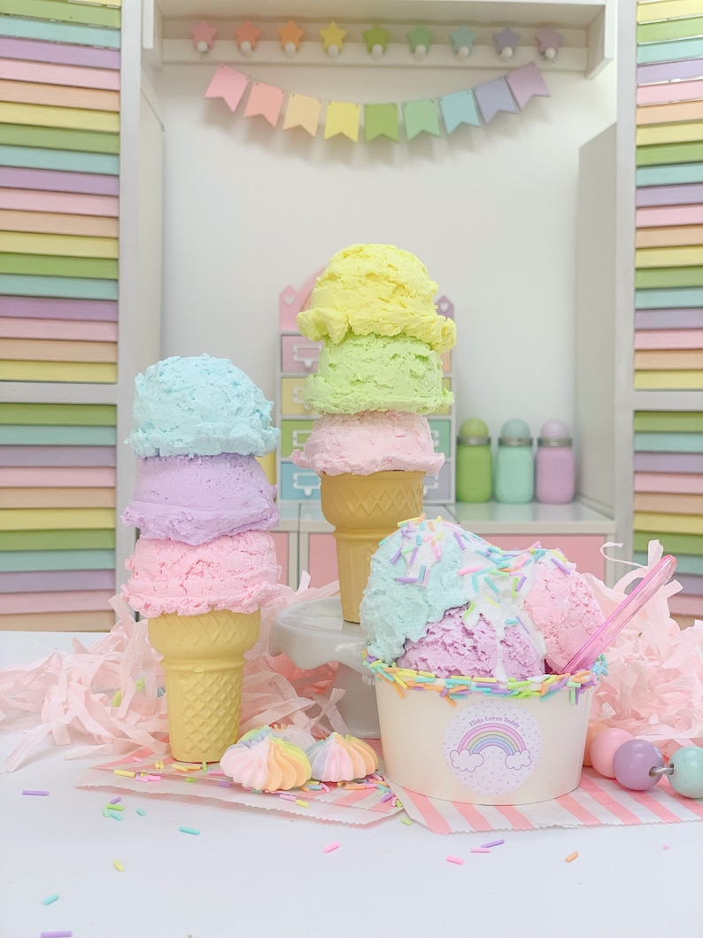 May include: Three pastel-colored ice cream cones with sprinkles and a bowl of ice cream with sprinkles. The ice cream is in a white bowl with a rainbow and the words "Kids Love Treats" on it. The ice cream is on a pink and white striped surface with a pink and white striped background.