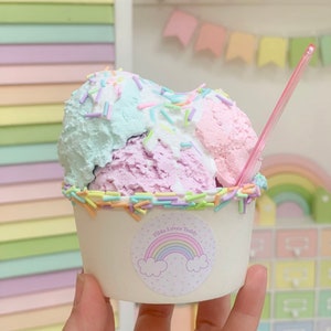 May include: A cup of ice cream with three scoops: pink, purple, and blue. The ice cream is topped with colorful sprinkles and a pink straw. The cup has a sticker with a rainbow and the text "Tilda Loves Teddy".