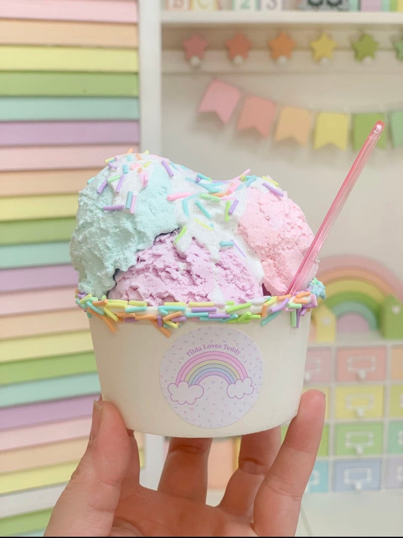 May include: A cup of ice cream with three scoops: pink, purple, and blue. The ice cream is topped with colorful sprinkles and a pink straw. The cup has a sticker with a rainbow and the text "Tilda Loves Teddy".