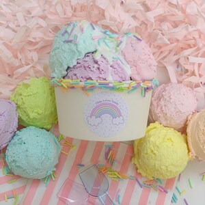 May include: A white ice cream cup with a rainbow sticker that says "Tilda Loves Teddy" and pastel-colored ice cream scoops. The ice cream is surrounded by pastel-colored sprinkles.