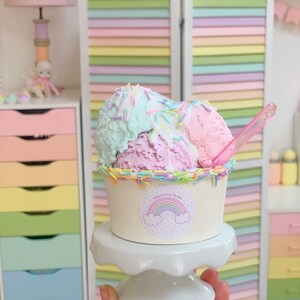 May include: A small white cup with a rainbow sticker that says "Tilda Loves Teddy" filled with three scoops of pastel-colored ice cream topped with colorful sprinkles. The ice cream is on a white cake stand.