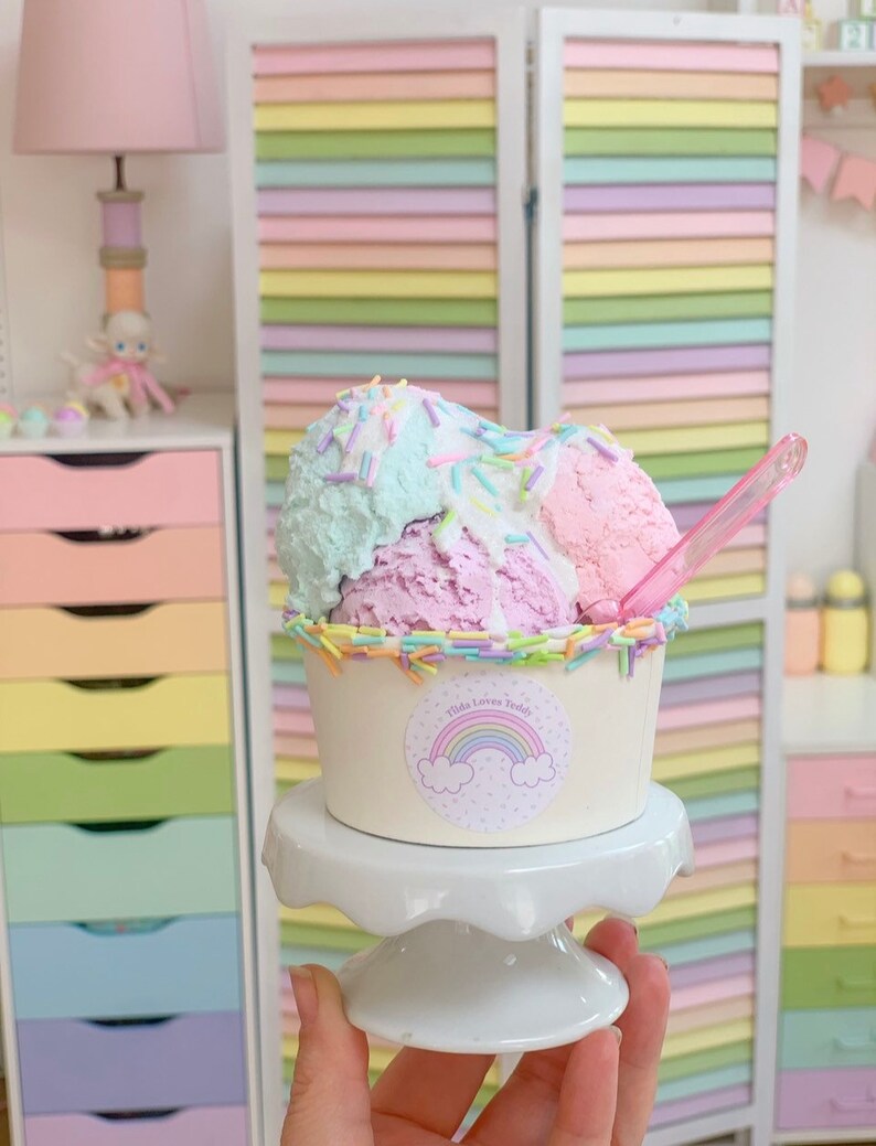 May include: A small white cup with a rainbow sticker that says "Tilda Loves Teddy" filled with three scoops of pastel-colored ice cream topped with colorful sprinkles. The ice cream is on a white cake stand.