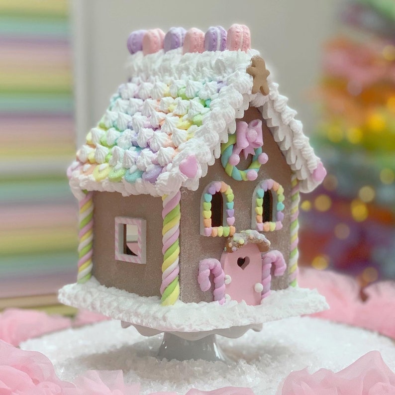 K&ouml;nnte beinhalten: Ein skurriles Lebkuchenhaus mit brauner Au&szlig;enseite, verziert mit pastellfarbenem Zuckerguss und S&uuml;&szlig;igkeiten. Das Dach hat einen Regenbogenverlauf, und das Haus steht auf einer wei&szlig;en Tortenplatte. Dekorative Details sind ein Lebkuchenmann und eine herzf&ouml;rmige rosa T&uuml;r.