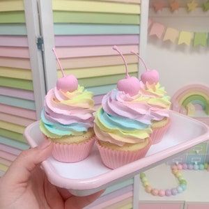 May include: Three pastel-coloured cupcakes with rainbow frosting and pink cherry toppings sit on a pink tray. The cupcakes are in pink paper cups. The background includes a pastel-coloured wall and decorative items.