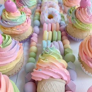Puede incluir: Cupcakes, donuts y un cono de helado de color pastel con chispas, dispuestos sobre una superficie blanca con una guirnalda de cuentas de color pastel.